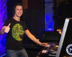 DJ mit Kopfhörern steht lächelnd hinter Pioneer-Equipment und zeigt Daumen hoch vor blau beleuchtetem Hintergrund