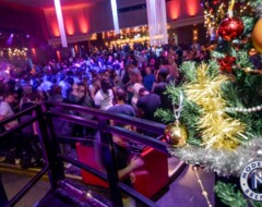 Weihnachtlich geschmückter DJ-Bereich mit Tannenbaum und Lichterketten über tanzender Menschenmenge in festlich beleuchteter Location