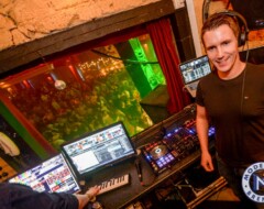 DJ an seinem Arbeitsplatz mit Mischpult, Laptops und Headset vor beleuchtetem Fenster mit tanzenden Gästen im Hintergrund
