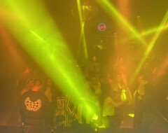 DJ-Pult im Vordergrund vor einer Bühne mit grün-orangenen Lichteffekten und tanzenden Menschen im Nebel