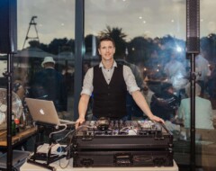 Hochzeits-DJ in Weste steht hinter DJ-Pult mit Mischpult und Laptop bei einer Abendveranstaltung mit Gästen im Hintergrund