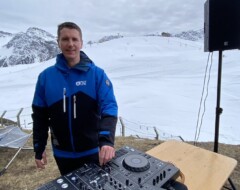 DJ an Mischpult in blauer Skijacke vor verschneiter Bergkulisse mit Skipiste im Hintergrund
