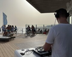 DJ mit Kopfhörern spielt auf einer Dachterrasse am Meer mit Serato-Controller für Gäste auf Sonnenliegen