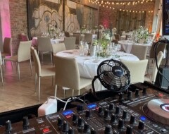 DJ-Equipment mit Mischpult im Vordergrund, dahinter festlich gedeckte Tische in einer Hochzeitslocation mit Backstein