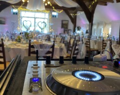 DJ-Pult mit leuchtenden Reglern im Vordergrund, dahinter festlich geschmückter Hochzeitssaal mit Lichterketten und Tischen