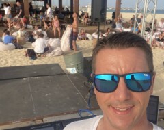 DJ mit Sonnenbrille am Mischpult bei Beach-Event mit Publikum auf Sand vor Strandbar