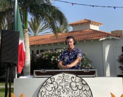 DJ mit Kopfhörern hinter Pult mit Totenkopf-Deko, Palmen und bunten Lichterketten im Hintergrund bei Outdoor-Event