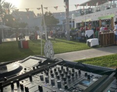 DJ-Equipment mit Mischpult im Vordergrund einer Outdoor-Veranstaltung mit Palmen, Wimpelketten und Gästen bei Sonnenschein