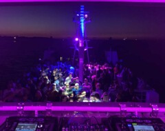 DJ-Pult mit Mischern und Plattenspielern vor tanzender Menschenmenge auf Boot bei Nacht mit lila Beleuchtung