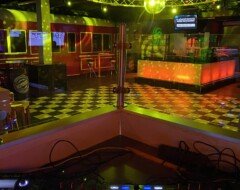 DJ-Pult mit leuchtenden Reglern vor einer Tanzfläche mit bunter Beleuchtung und Bar im Hintergrund